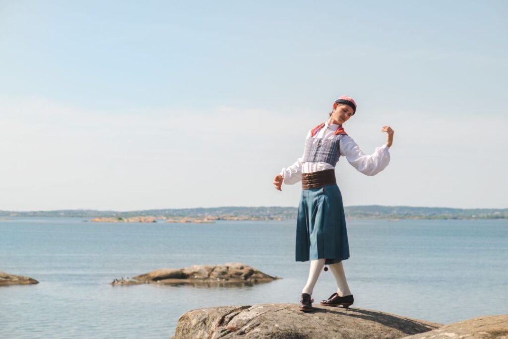 Fredy Clue står på en klippa vid havet, iförd en folkdräkt med vit skjorta, väst och blå kjol.