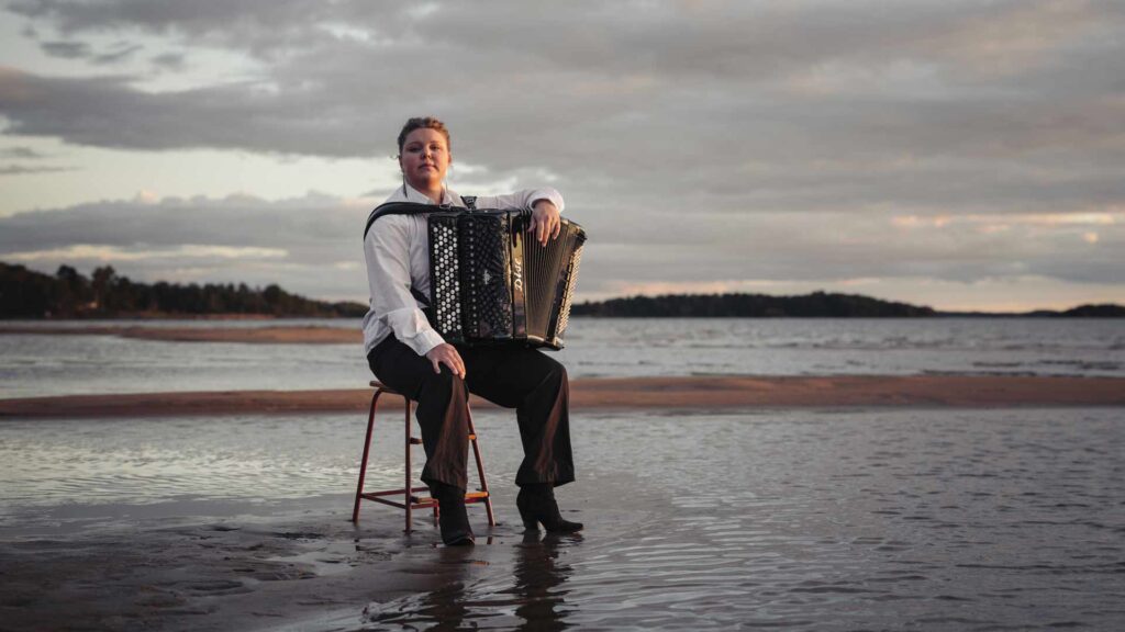 En musiker med dragspel, Larisa Ljungkrona-Mönttinen, sitter på en stol i vattenbrynet vid havet