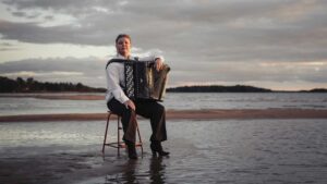 En musiker med dragspel, Larisa Ljungkrona-Mönttinen, sitter på en stol i vattenbrynet vid havet