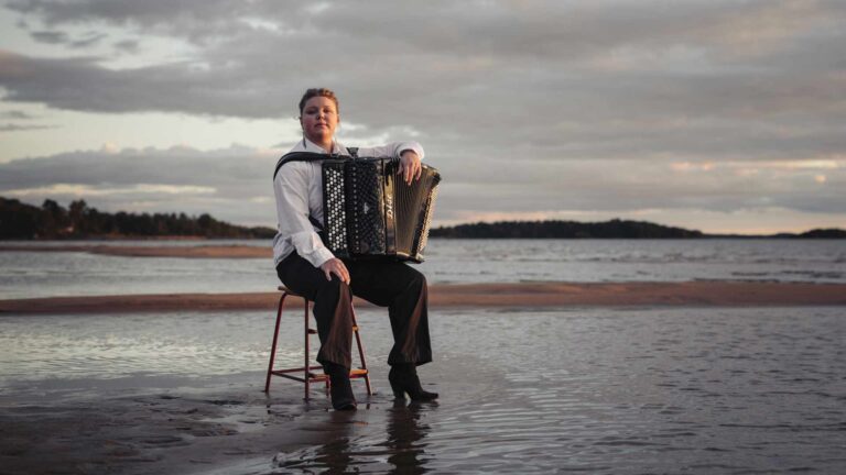 En musiker med dragspel, Larisa Ljungkrona-Mönttinen, sitter på en stol i vattenbrynet vid havet