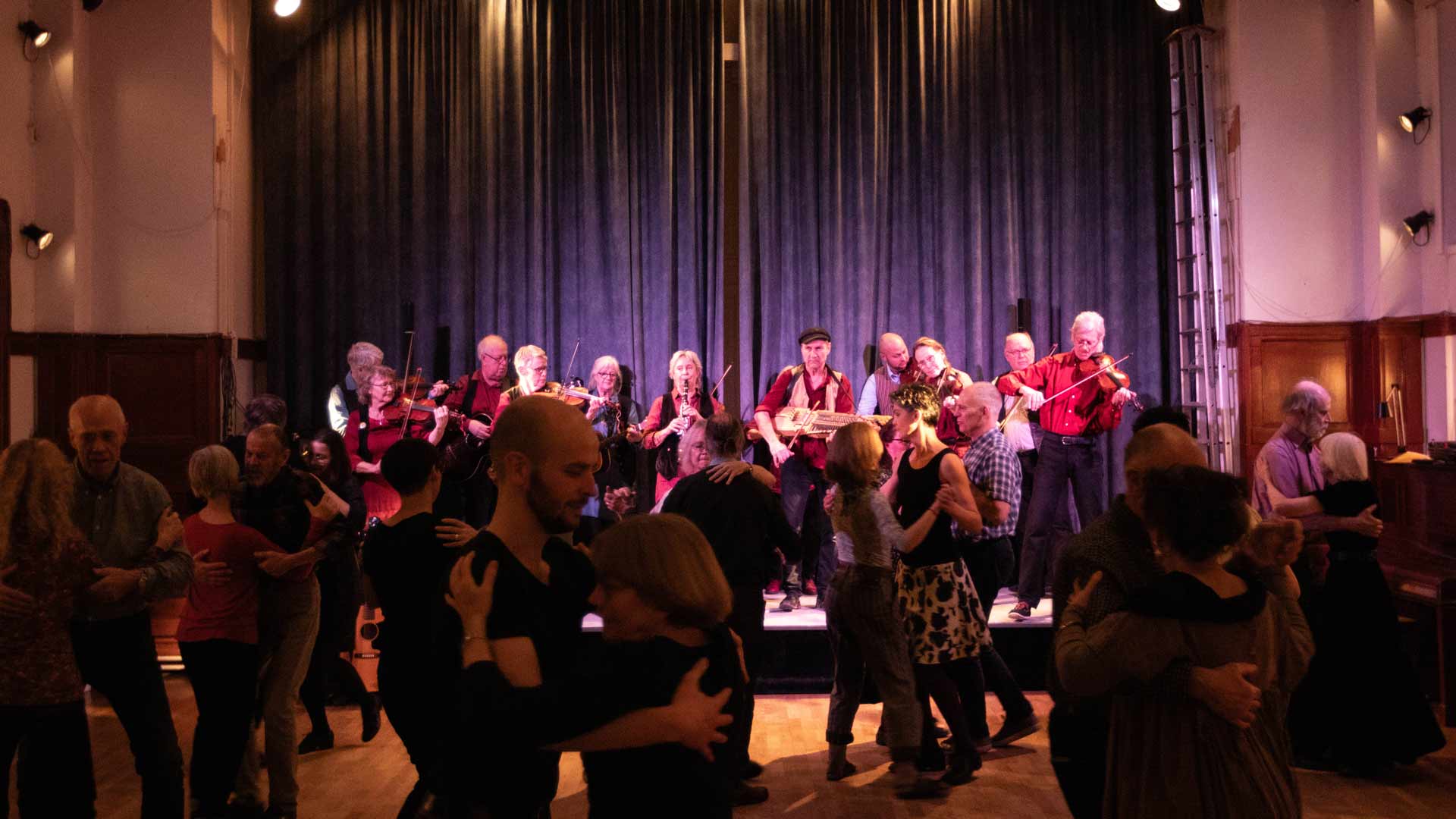 Dansstuga med spelmän på scenen och dansare i alla åldrar framför